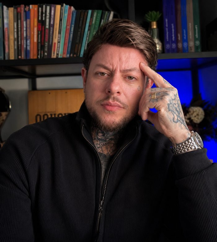 Greg Pàz em seu escritório, apoiado na mão, com estante de livros ao fundo.